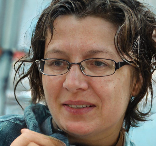 Close-up of a woman with short curly brown hair and glasses, smiling slightly at the camera